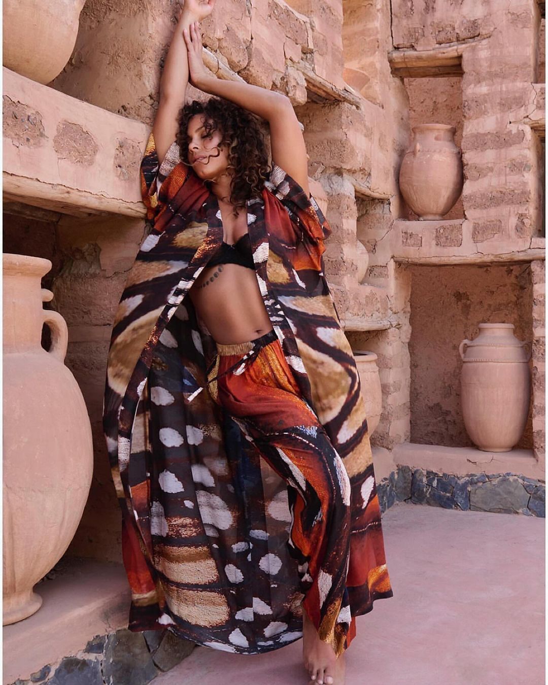 A woman in butterfly-patterned clothing poses in a desert environment with clay jugs, showcasing a blend of fashion and natural beauty.