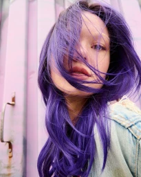 Close-up portrait of a woman with vibrant purple hair blowing across her face, wearing a denim jacket against a pink wall.