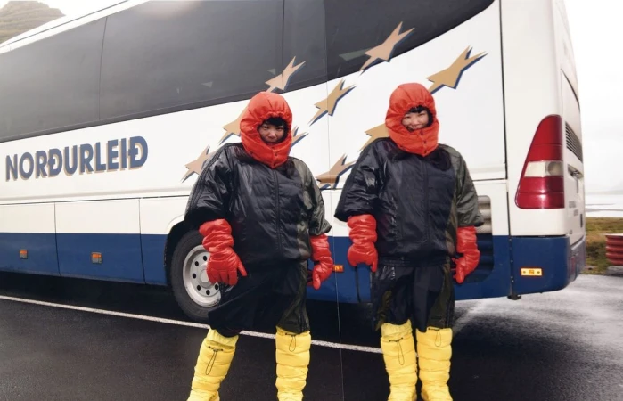 Two people in matching protective suits stand in front of a bus in an outdoor, overcast setting. The people face the camera.