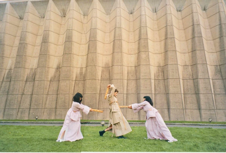 Three women in pink dresses engage in a playful baguette fight on a grassy field in front of a large concrete building.