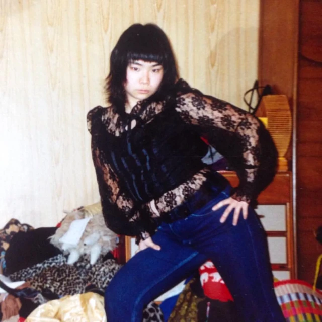 An Asian woman poses in a black lace top and blue jeans, set against a simple backdrop with a pile of fabrics in the foreground.