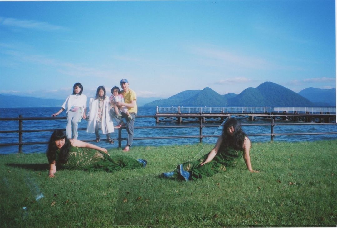 Family portrait near a lake with mountains in the distance; two women in green dresses pose in the foreground, with a clear sky.