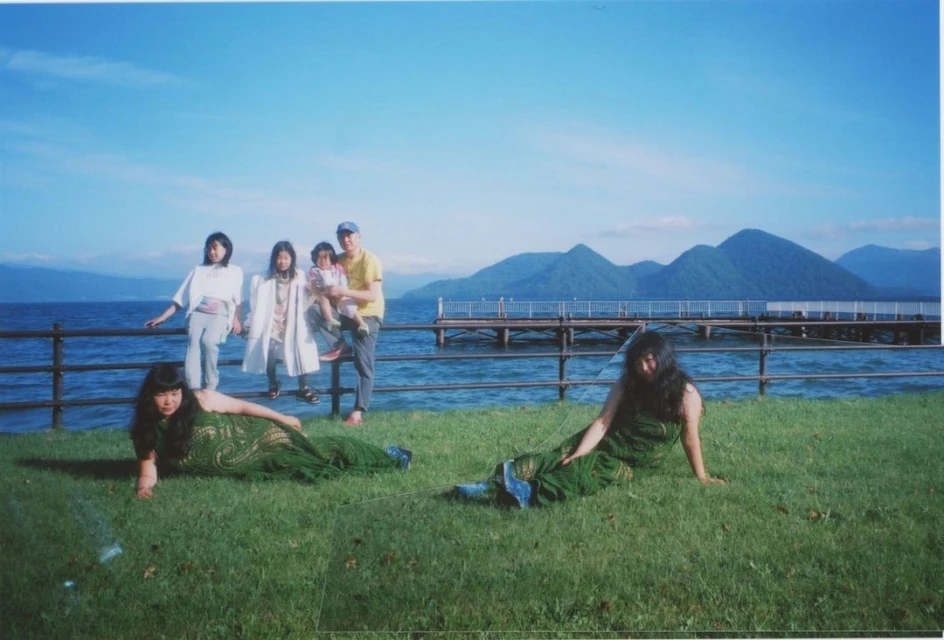 Family portrait near a lake with mountains in the distance; two women in green dresses pose in the foreground, with a clear sky.
