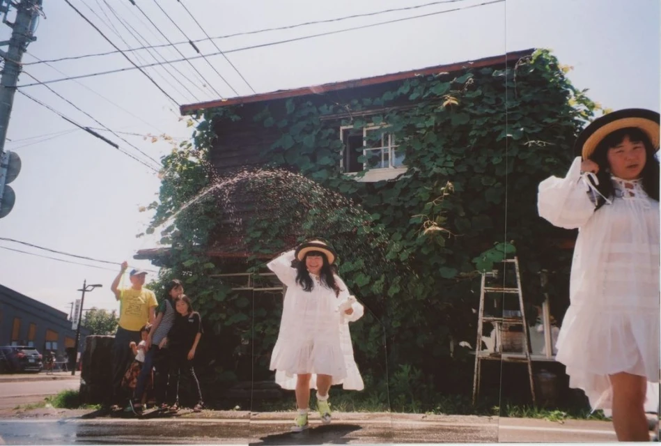 A group of people are outdoors on a sunny day in an urban environment with a building covered in ivy in the background.