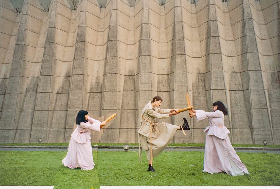 Three people in vintage costumes engage in a playful sword fight with baguettes on green grass in front of a textured concrete wall.