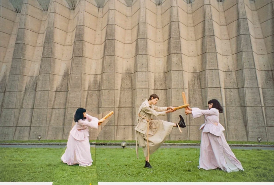 Three people in vintage costumes engage in a playful sword fight with baguettes on green grass in front of a textured concrete wall.