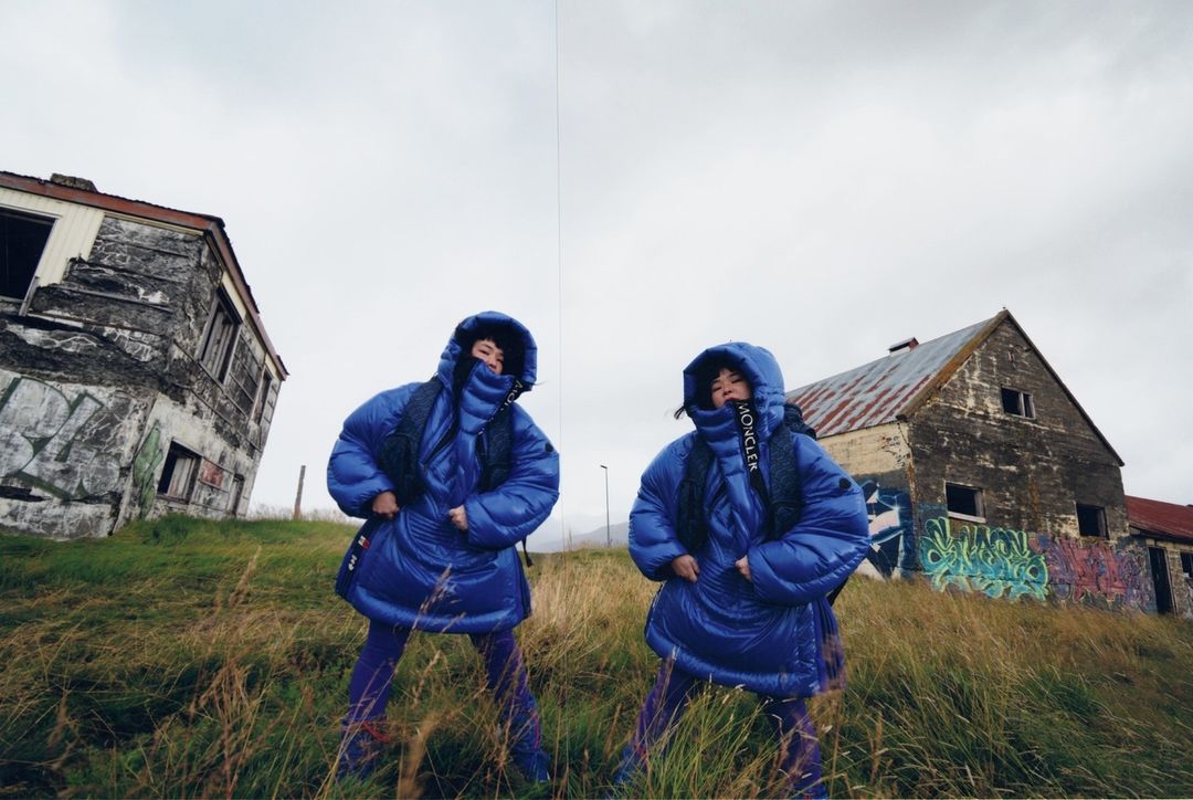 Two women in matching blue hooded puffer jackets pose in a grassy field near dilapidated buildings under an overcast sky.
