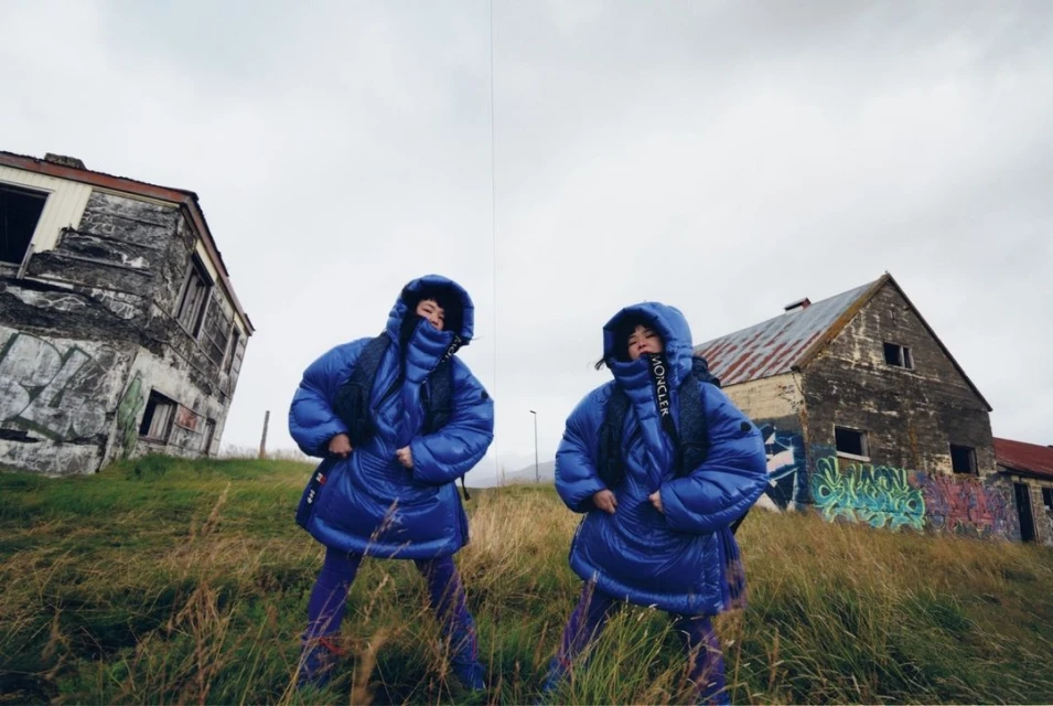 Two women in matching blue hooded puffer jackets pose in a grassy field near dilapidated buildings under an overcast sky.