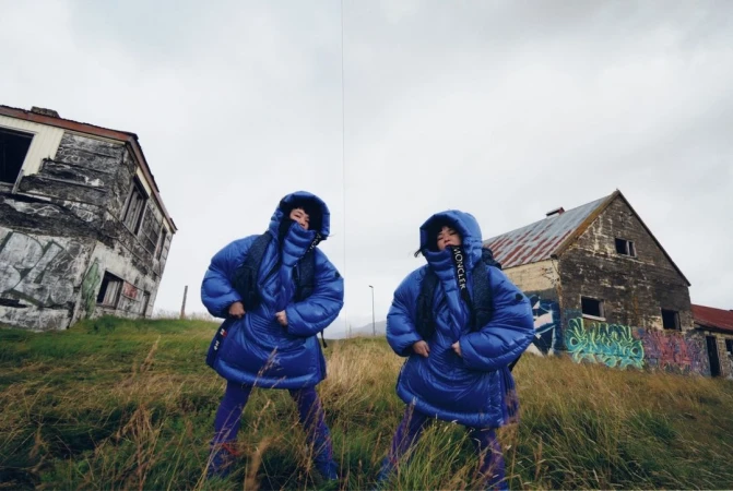 Two women in matching blue hooded puffer jackets pose in a grassy field near dilapidated buildings under an overcast sky.
