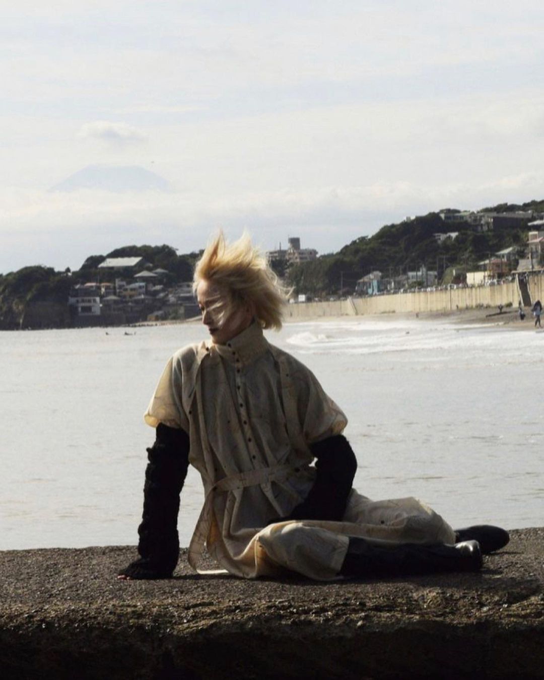 Woman with stylized makeup sitting on a concrete structure with the ocean, shoreline, and a distant mountain in the background.