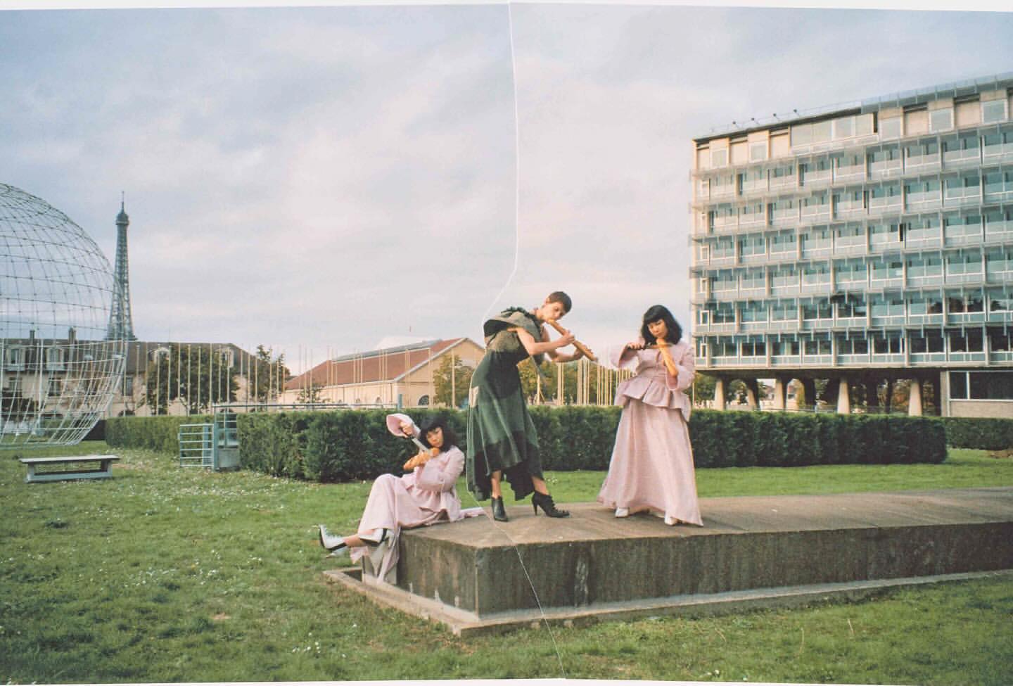 Three women pose outdoors in pink and green vintage gowns with props; lawn, modern architecture, and Eiffel Tower in the background.
