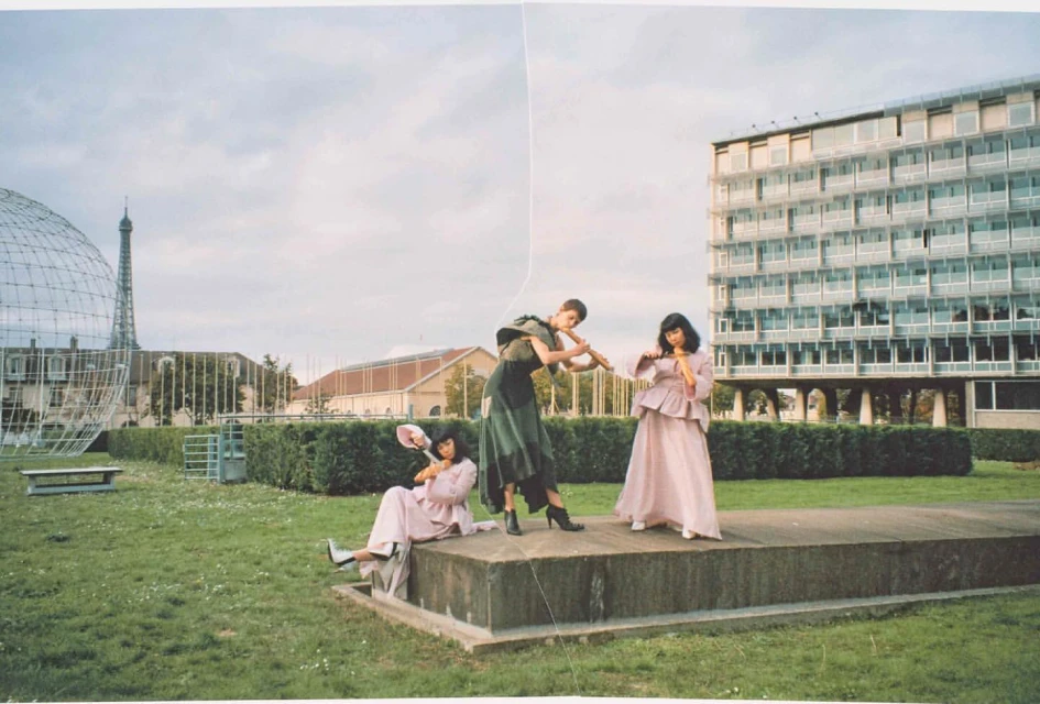 Three women pose outdoors in pink and green vintage gowns with props; lawn, modern architecture, and Eiffel Tower in the background.