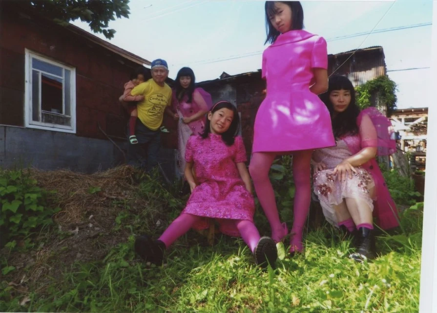 Group of Asian women and a man with a child pose outdoors in pink attire near a weathered building on a sunny day.