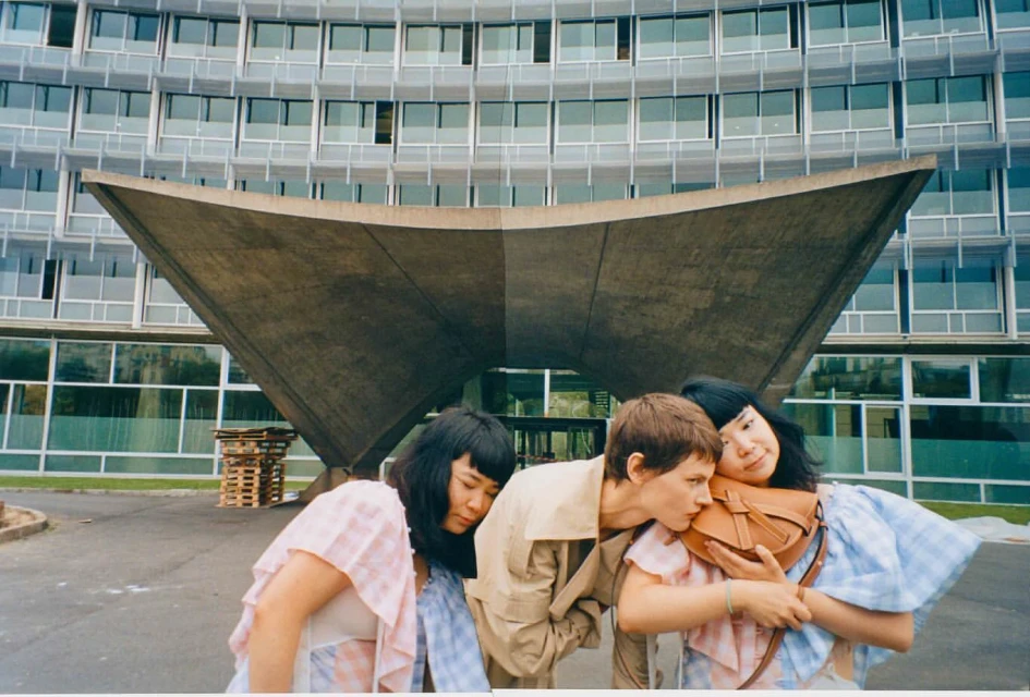 Three models pose outdoors in front of a modern building. One model hugs a brown handbag. They wear casual neutral and blue-and-white-checkered outfits.