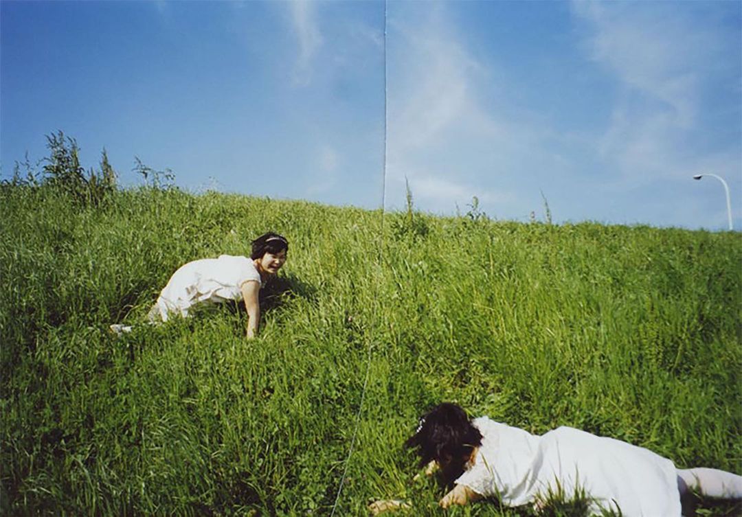 Two young women in white dresses climb a grassy hill under a bright blue sky with scattered clouds, creating a whimsical and serene scene.