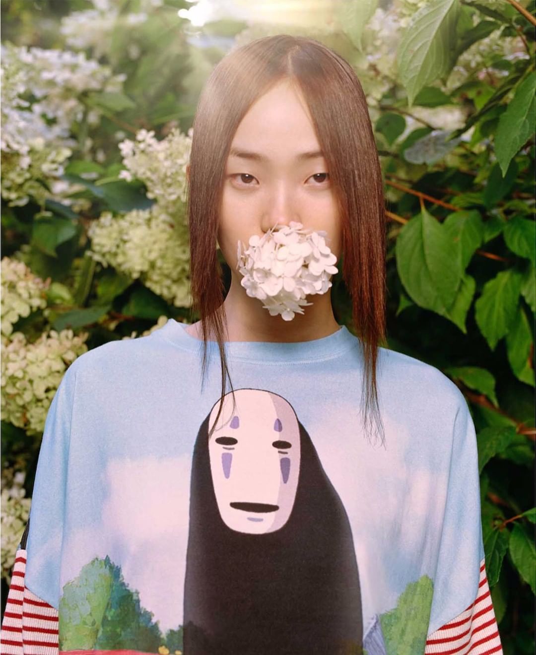 A woman with straight brown hair and a T-shirt featuring a cartoon character holds a white flower to her mouth outdoors.