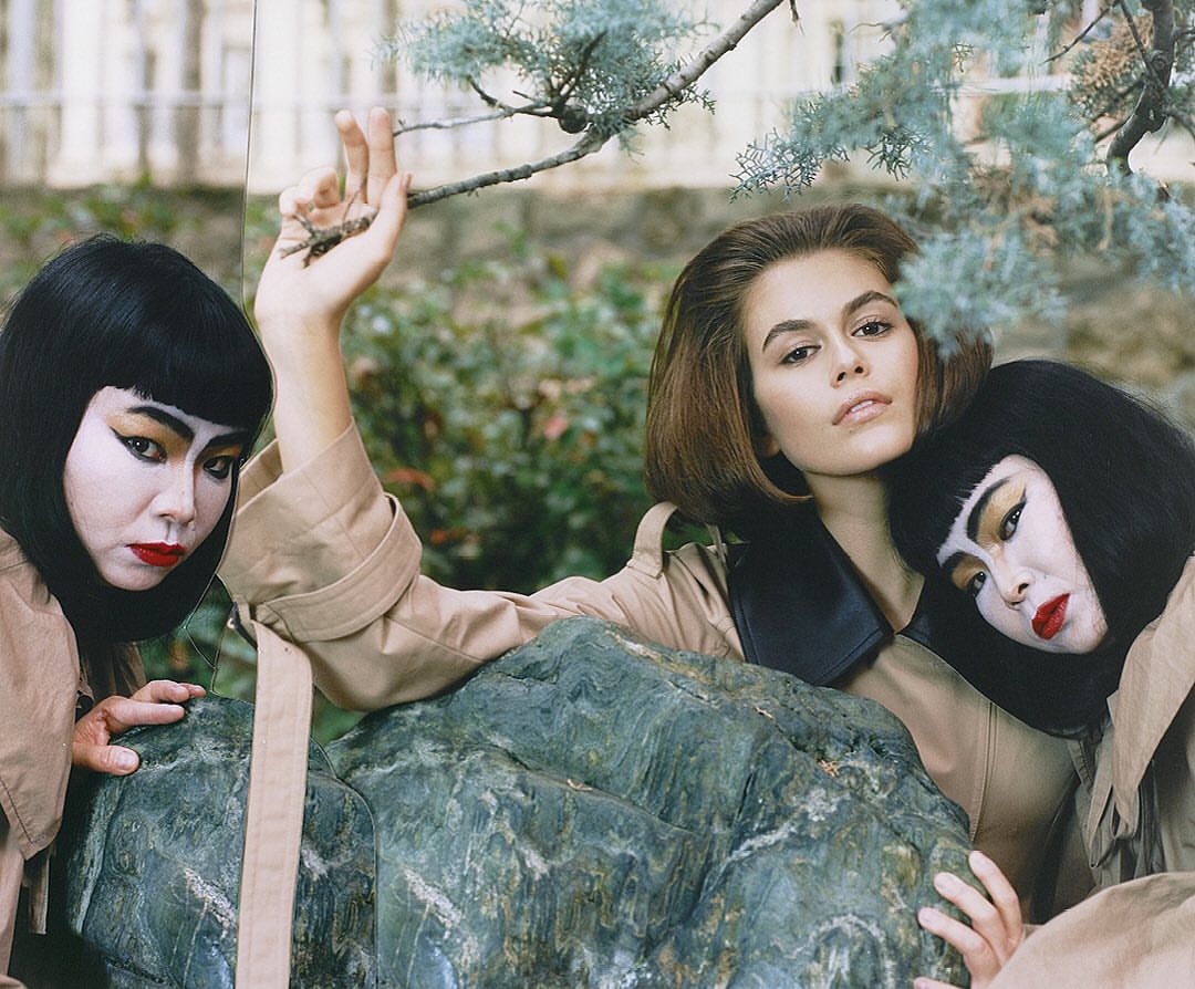 Fashion editorial shot featuring a model with short brown hair and two models in geisha-style makeup, posing outdoors in front of greenery.