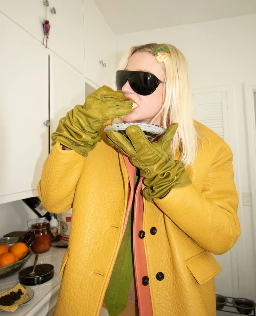 Woman in yellow coat and green gloves eats from plate in kitchen. Wearing sunglasses and flower hair clip in bright, indoor setting.