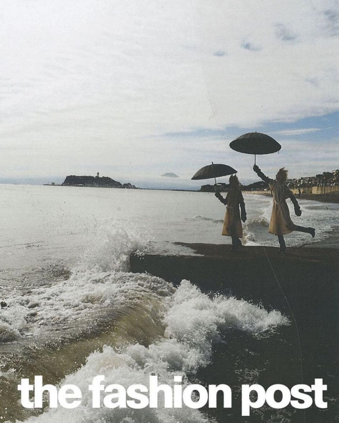Two women in dresses with umbrellas stand on a rocky shoreline near the ocean and a distant island under a cloudy sky. Fashion editorial.