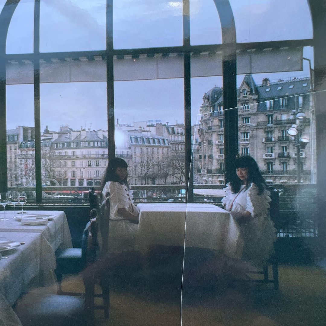 Two adult females sitting at a table in a restaurant, with a view of city buildings through a large arched window in the background.