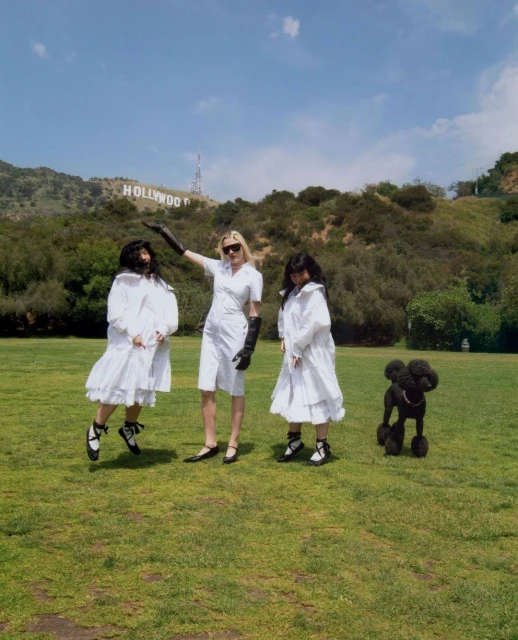 Three women in white dresses pose with a poodle in front of the Hollywood sign, creating a whimsical fashion editorial.