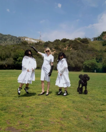 Three women in white dresses pose with a poodle in front of the Hollywood sign, creating a whimsical fashion editorial.