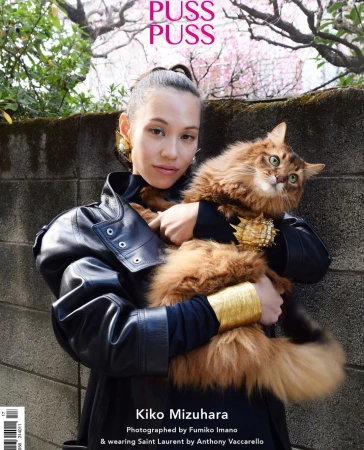 Fashion editorial featuring a woman in a black jacket holding a fluffy cat with gold accessories, outdoor setting with flowering trees.