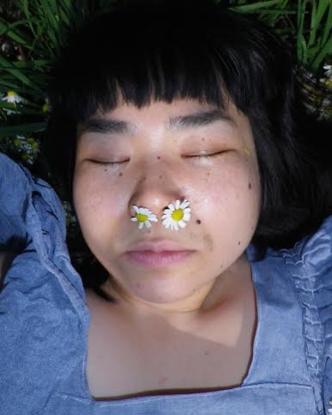 Close-up of an Asian woman with closed eyes lying in green grass and daisies, two daisy heads playfully placed on her nose.
