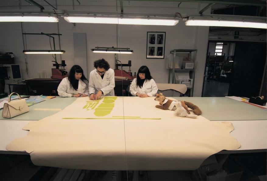 Three people in lab coats work at a table with a handbag, templates, material, and a fox toy in a bright indoor setting.