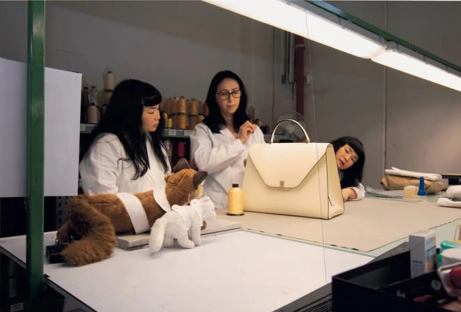 Two women in lab coats inspect a cream-colored handbag on a table in a brightly lit workshop, next to plush toy animals and spools of thread.