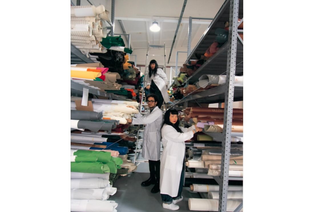Three people in lab coats stand among shelves stocked with rolls of fabric in an indoor industrial setting with bright, even lighting.