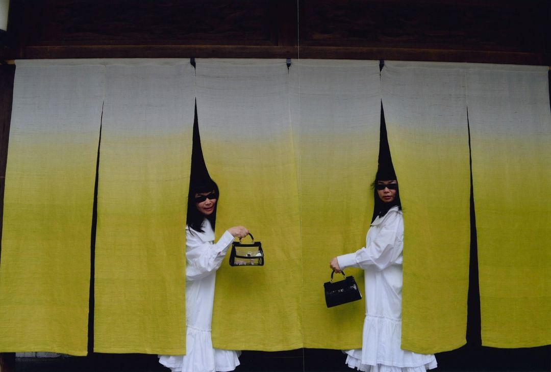 Two adult women stand behind an opening in a yellow-white curtain, each holding a black handbag, wearing sunglasses and cone-shaped headwear.