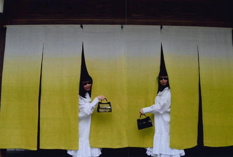 Two adult women stand behind an opening in a yellow-white curtain, each holding a black handbag, wearing sunglasses and cone-shaped headwear.