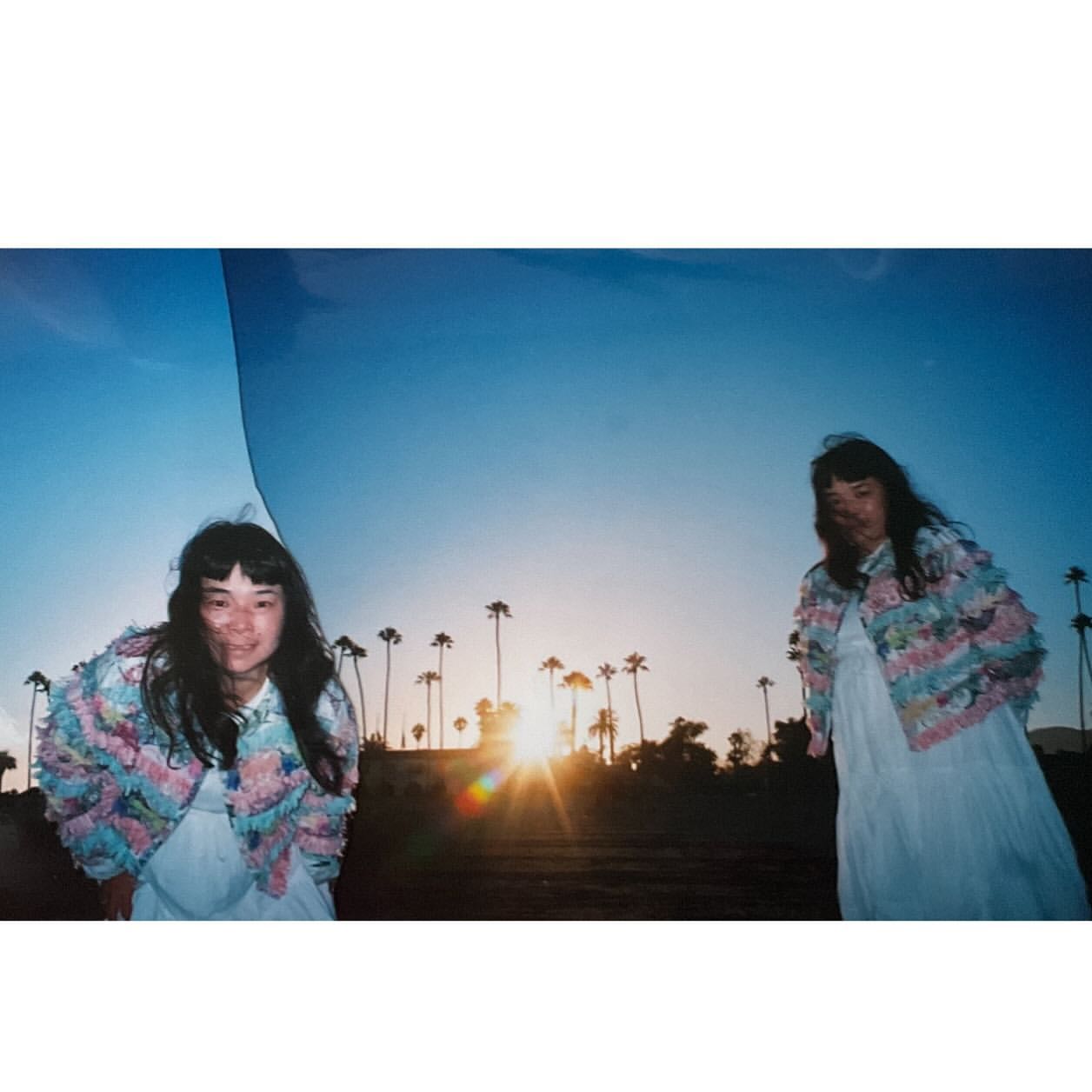 Two women pose outdoors against a desert sunset and palm trees, wearing jackets with fringe and light-colored dresses.