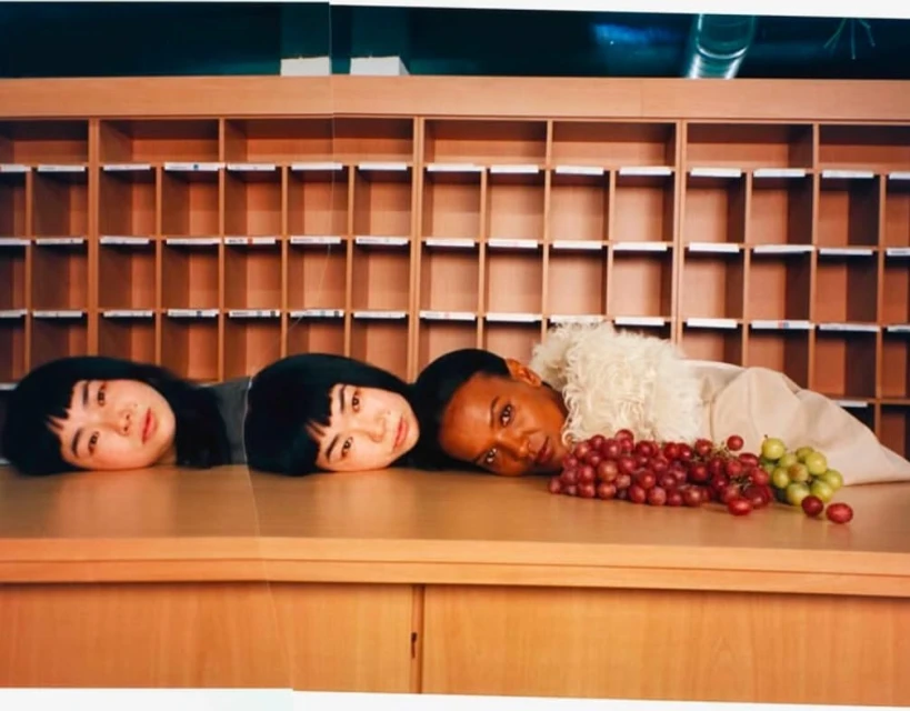 Three women with dark hair lying on a tan counter next to bunches of red and green grapes, in front of a wall of cubbies.