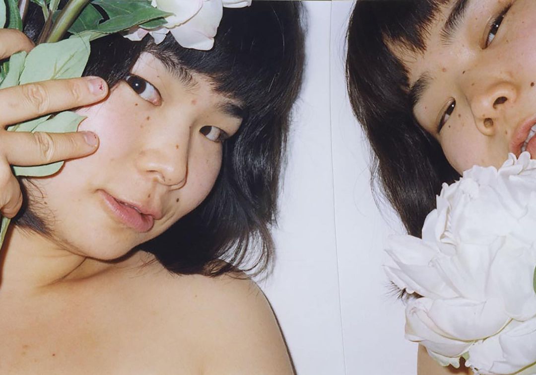 Close-up studio shot of two young Asian women with dark hair and flowers, one making a peace sign, against a white backdrop.