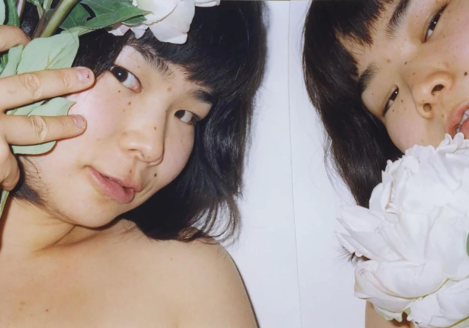 Close-up studio shot of two young Asian women with dark hair and flowers, one making a peace sign, against a white backdrop.
