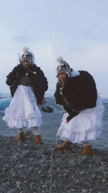 Two women wearing winter hats and coats pose near icebergs at Jokulsarlon glacier lagoon in Iceland with a bright sky and water.