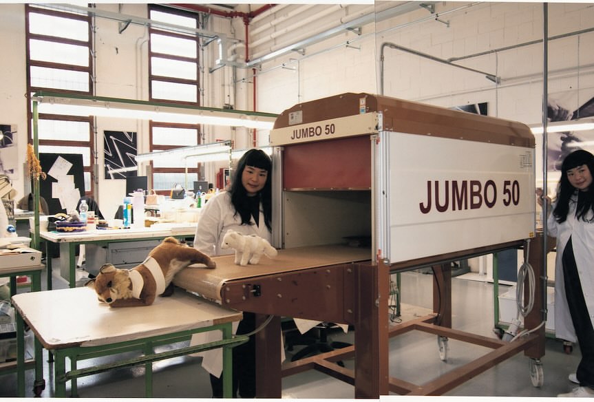 Two women in lab coats stand next to a machine, with a plush fox and polar bear toy on a table in an indoor laboratory setting.