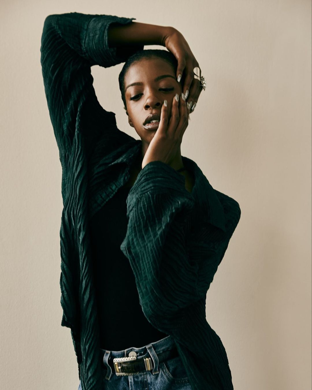 Portrait of a Black woman with hands near her face, wearing a black top and a wrinkled dark green coat in a neutral indoor setting.