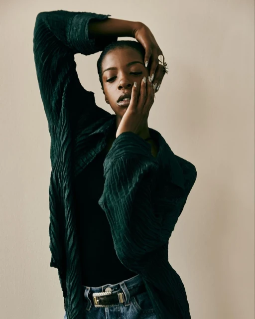 Portrait of a Black woman with hands near her face, wearing a black top and a wrinkled dark green coat in a neutral indoor setting.