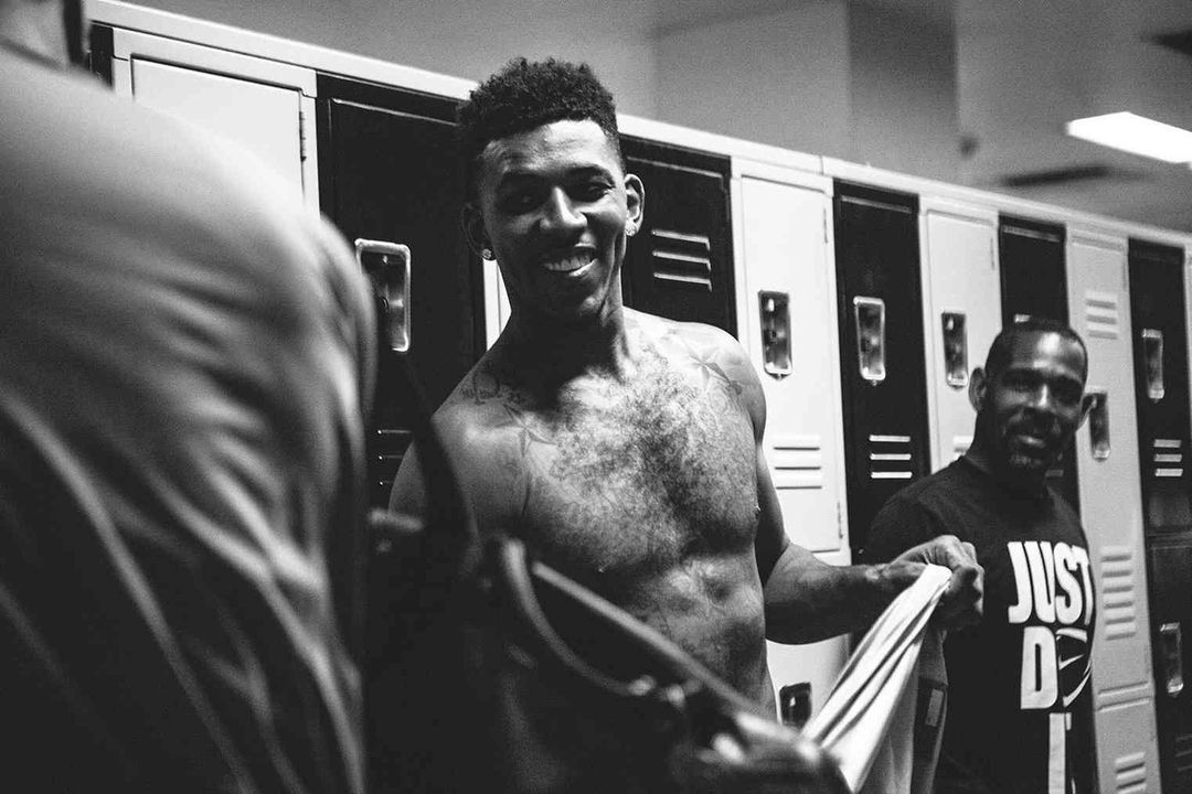 Black and white shot of a smiling man in a locker room, standing with a towel, along with two other men in the background.
