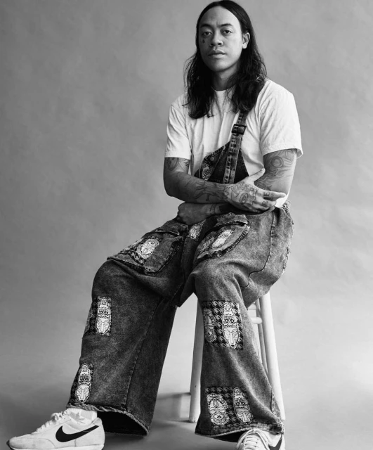 Black-and-white portrait of a man with long hair and tattoos wearing overalls sitting on a stool in a photo studio setting.