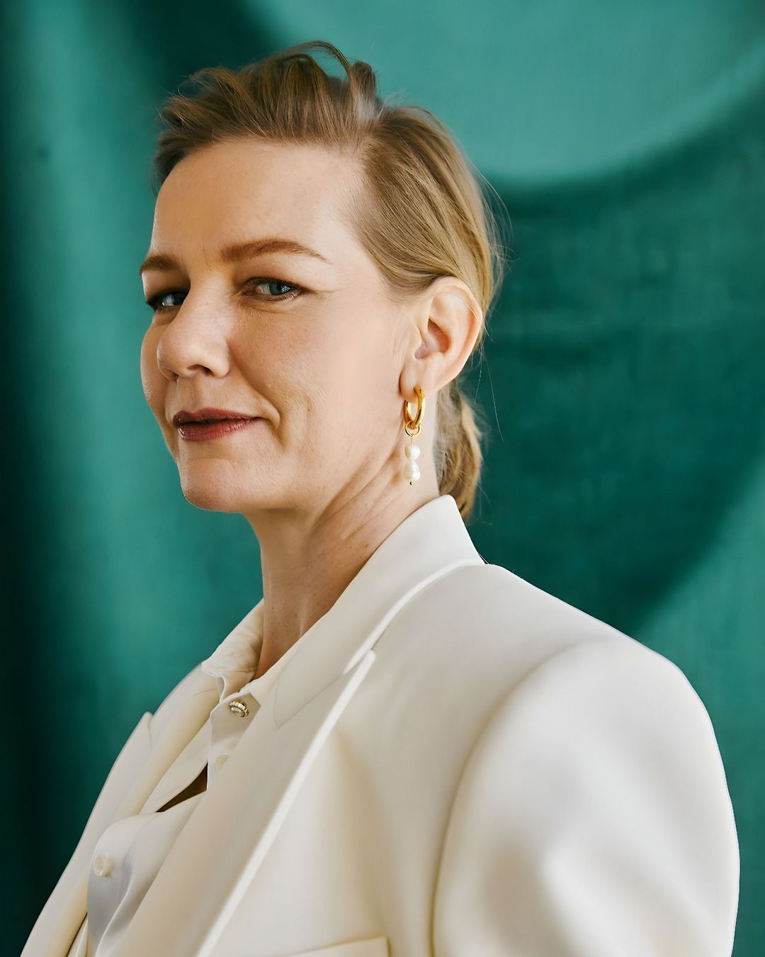 Portrait of a blonde woman wearing a white blazer and gold hoop earrings against a textured green background.
