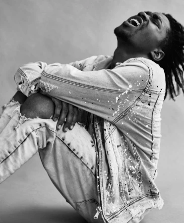 Dramatic black and white portrait of a young Black man in a paint-splattered denim jacket, expressing emotion with his mouth open.