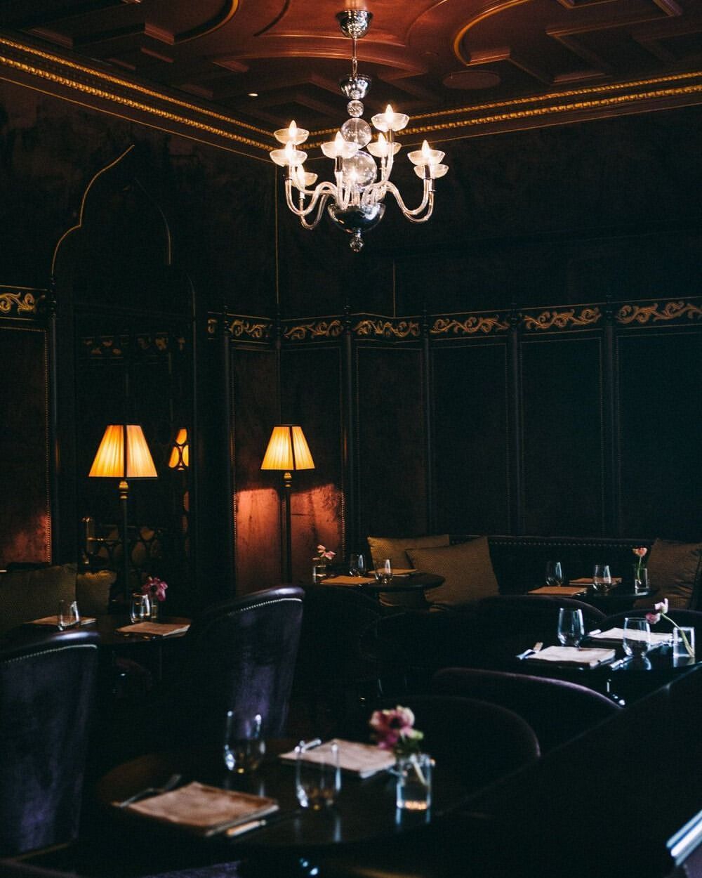 Elegant interior with chandelier, tables set with glassware and flowers, plush chairs, and warm lamp lighting in a sophisticated ambience.
