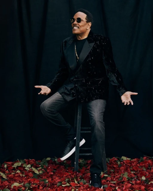 Portrait of a smiling adult male with sunglasses, seated on a ladder, wearing a black sequined jacket, jeans, and black sneakers.