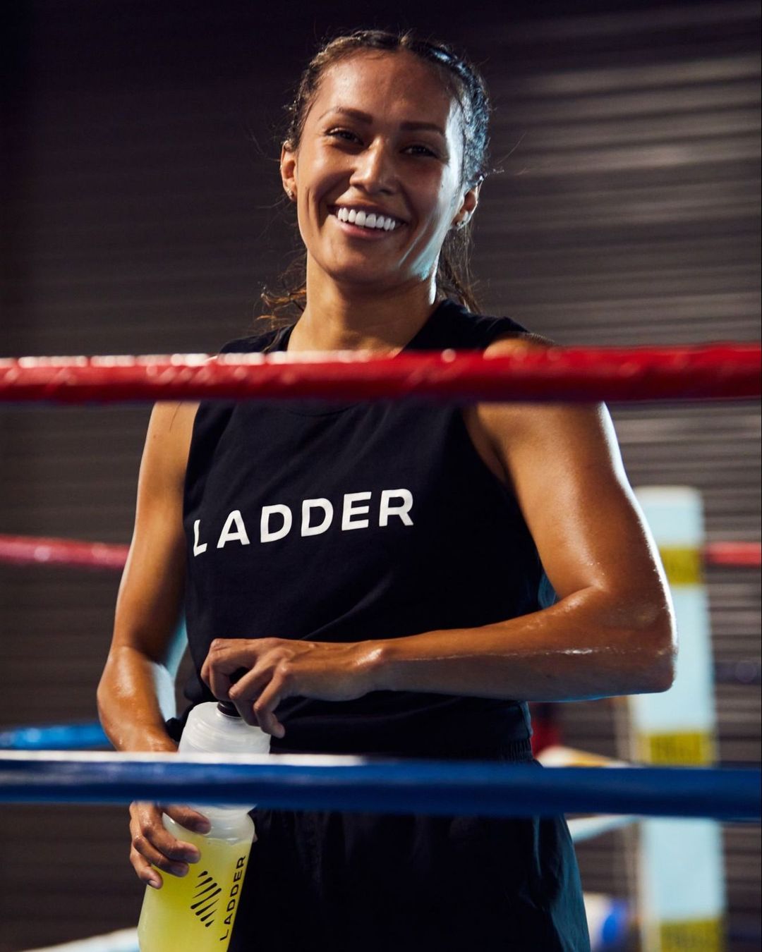 Smiling woman in fitness attire, holding a water bottle after exercise in a boxing ring, promoting health and well-being.