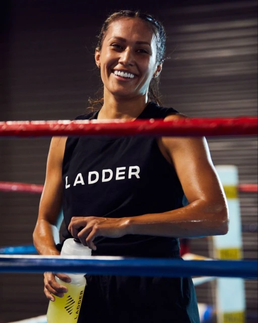 Smiling woman in fitness attire, holding a water bottle after exercise in a boxing ring, promoting health and well-being.