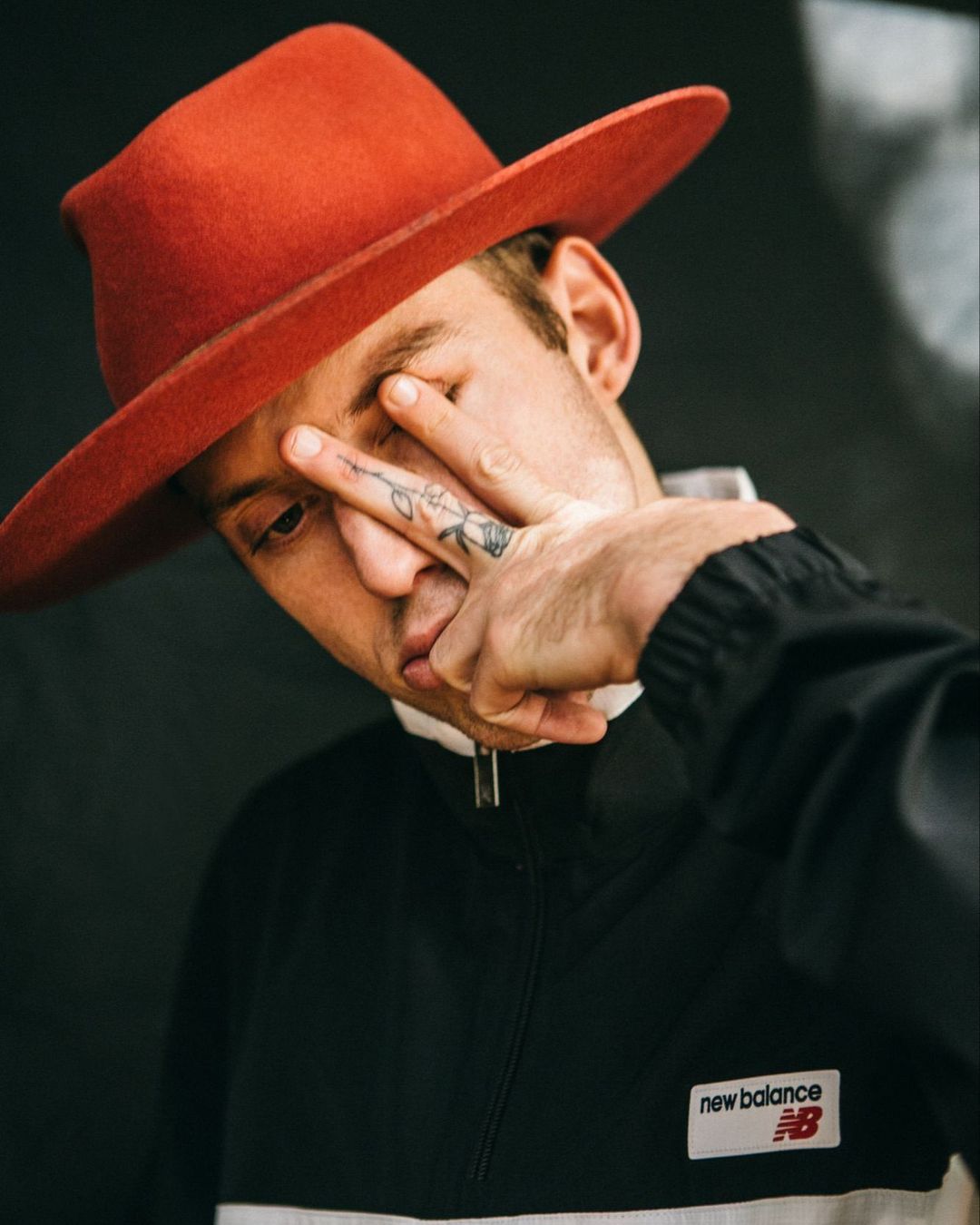 Close-up of a person wearing an orange hat and a black and white jacket, posing with fingers near the eye against a dark background.